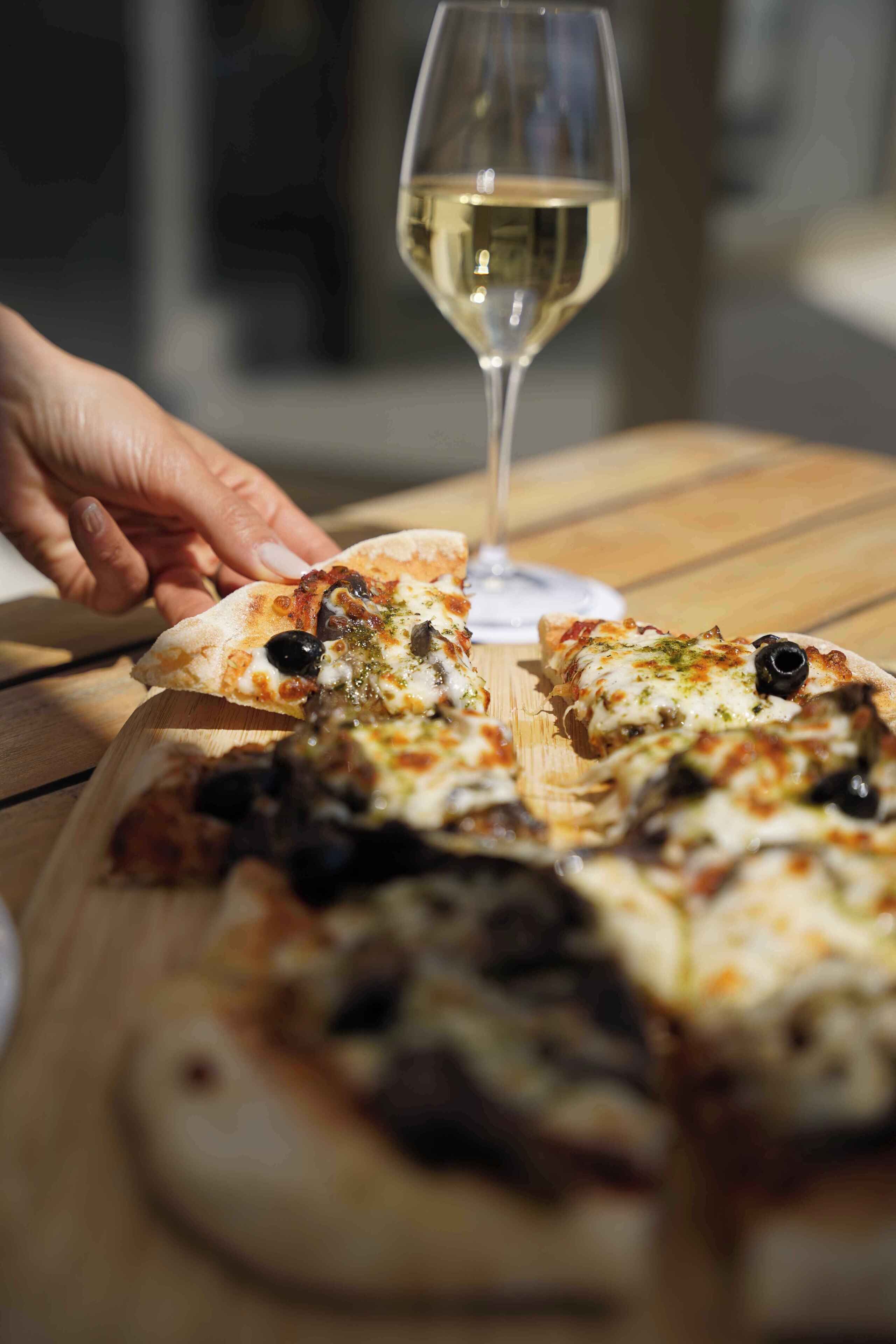 Oliiveilla päällystetty pizza ja lasillinen valkoviiniä puupöydällä.