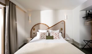 Une chambre d’hôtel comprenant un lit, une table de chevet, une commode, une lampe fixée au mur et une fenêtre avec des rideaux.