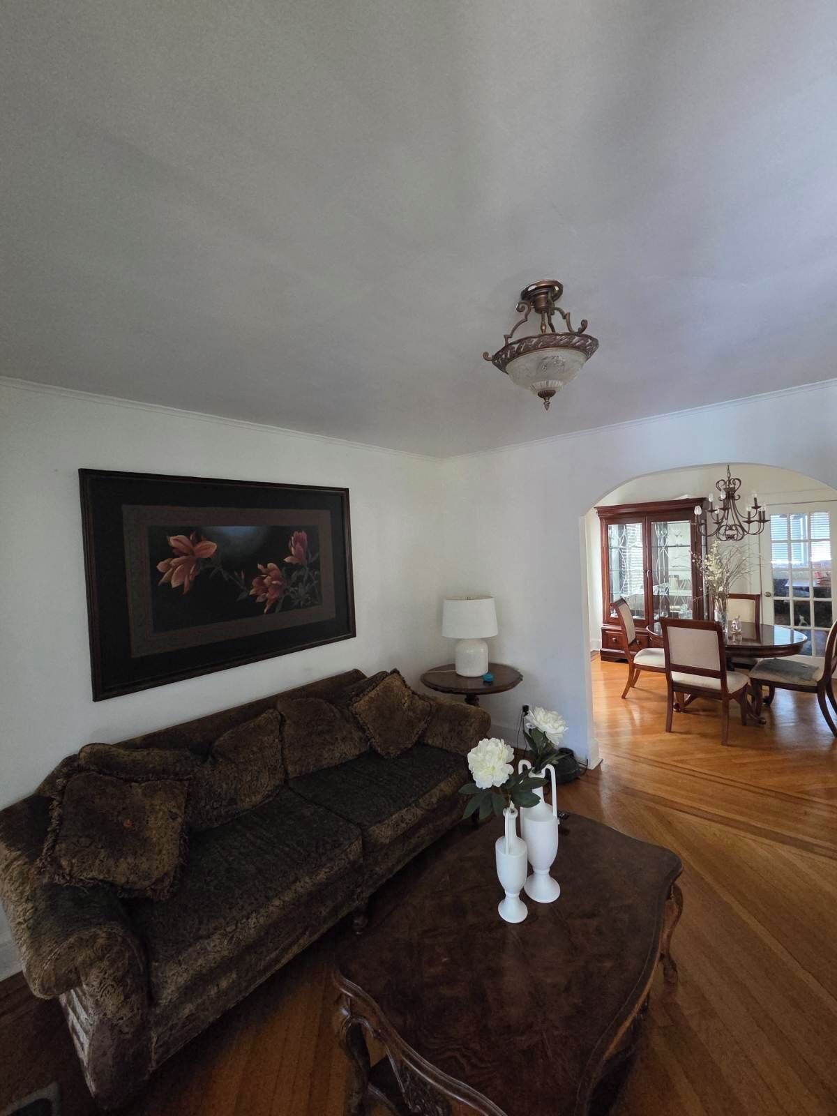 Uma sala de estar com um sofá marrom escuro, uma mesa de centro com vasos brancos, um quadro com flores na parede e uma área de jantar com mobiliário de madeira.