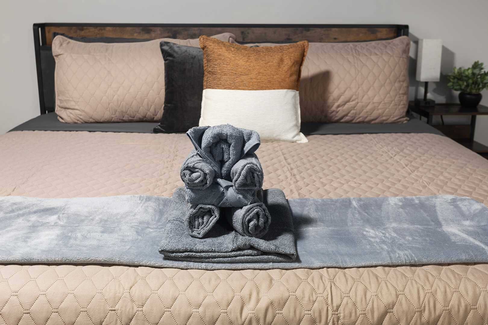 Una cama bien hecha con un cabecero acolchado, una toalla enrollada y una pequeña planta en maceta sobre la mesita de noche.