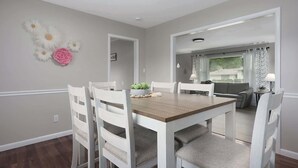 A dining area with a wooden table and white chairs, a small plant centerpiece, and a wall with decorative flowers. - Family Favorite_Arcades_ KingBeds_ Sleeps 10_ BBQ (Shawnee)