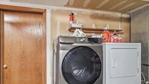 Een wasruimte met een wasmachine en droger, een houten deur en een plank met schoonmaakspullen.