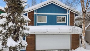 Une maison à deux étages, à la façade bleue, avec un garage et un arbre enneigé devant.