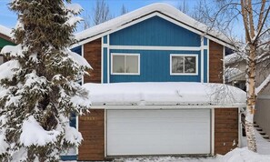 Ein zweistöckiges Haus mit blauer Fassade, einer Garage und einem schneebedeckten Baum davor.
