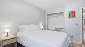Une chambre à coucher avec un lit, des tables de chevet, une fenêtre et un tableau abstrait aux couleurs vives accroché au mur.