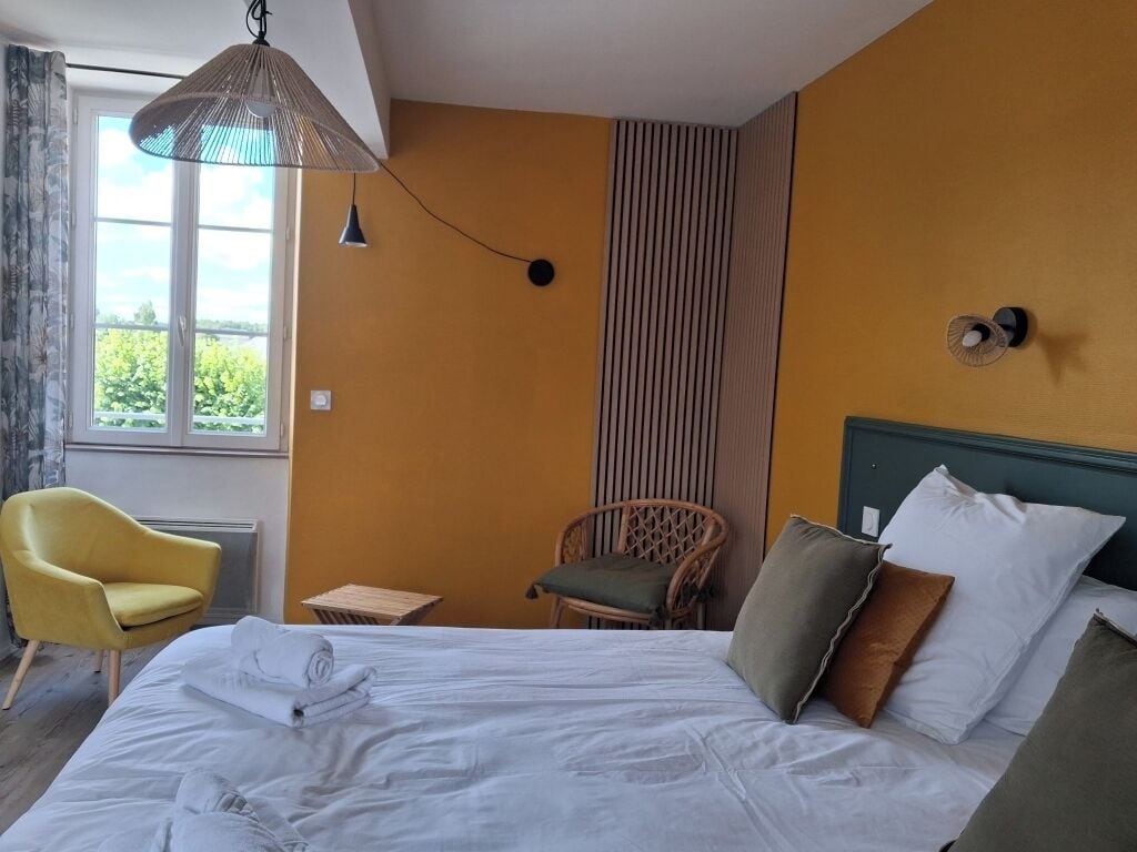 Habitación de hotel con cama, un sillón amarillo, una silla de mimbre, una mesita y una ventana con cortinas.
