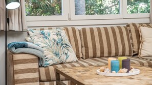 Un sofá a rayas con un cojín azul y blanco, una mesa de centro de madera con velas y conchas marinas, y una ventana con cortinas.
