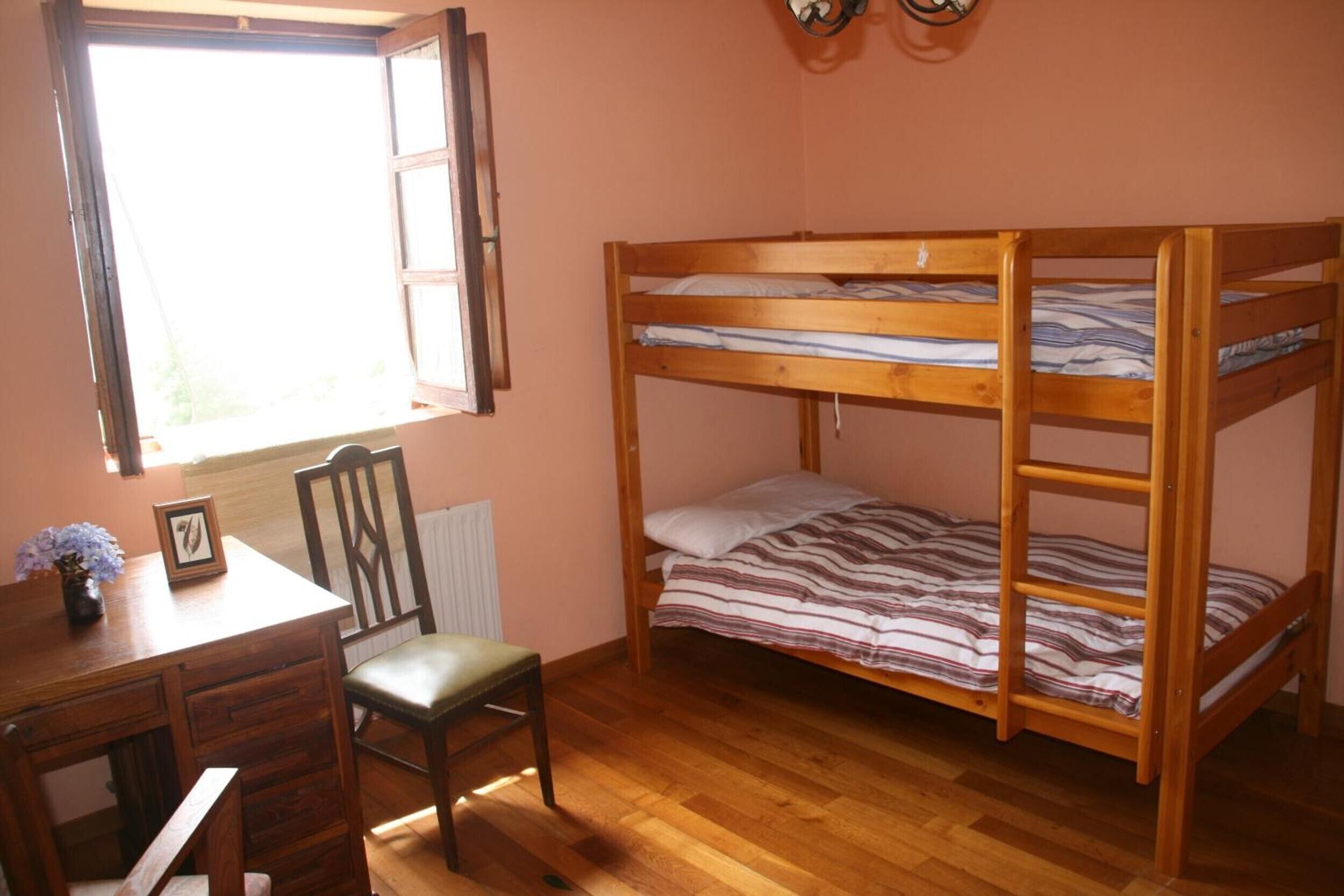A bunk bed with a wooden frame, a desk with drawers, a chair, a radiator, and a window with open shutters.