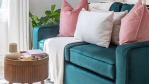 A teal sofa with multiple pillows, a small wooden side table with a vase and books, and a green plant in the background. - Cosy 2BR in Marylebone Near Baker Street (London)