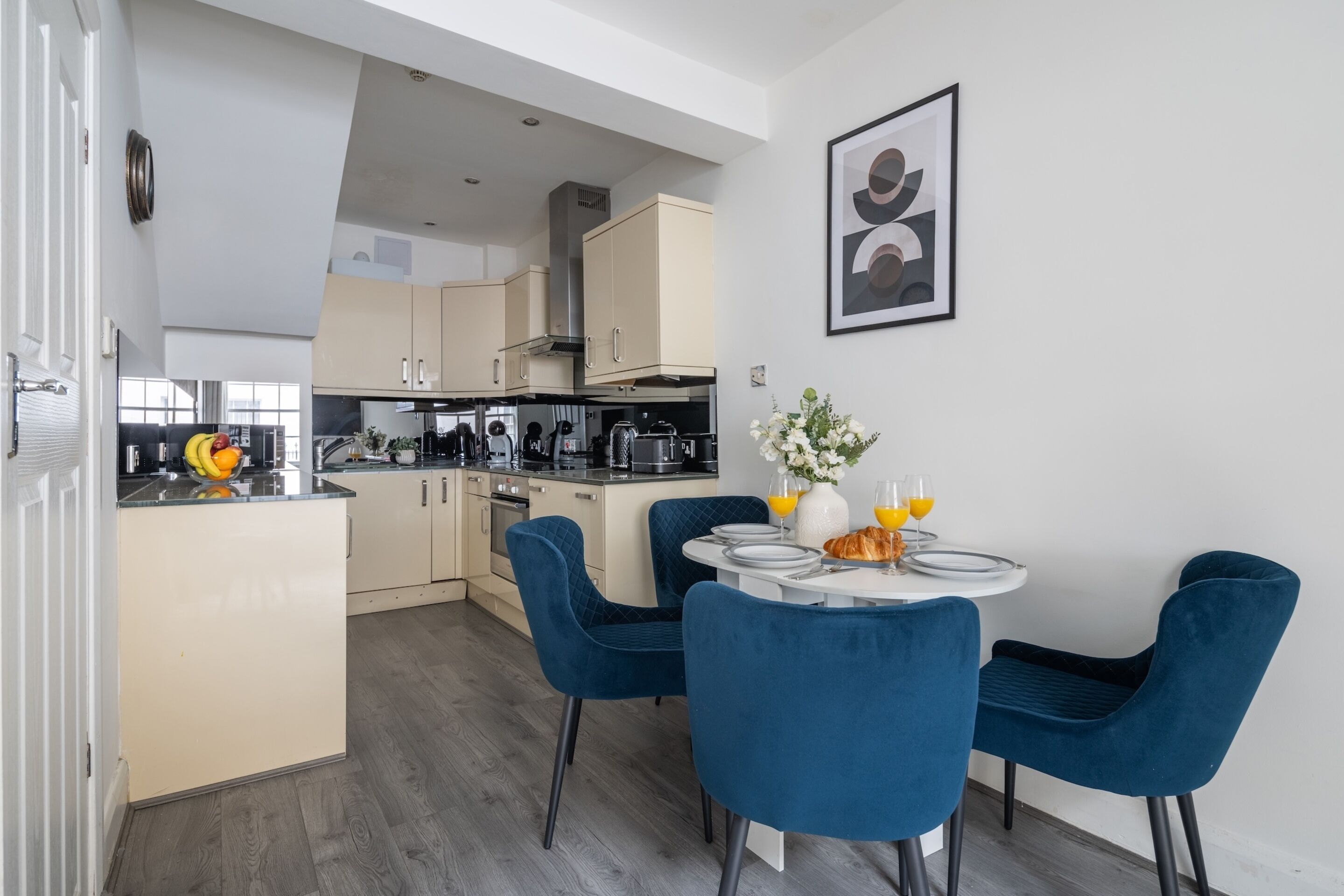 A modern kitchen with a dining area featuring blue chairs and a table set with plates, a vase with flowers, and orange juice glasses.