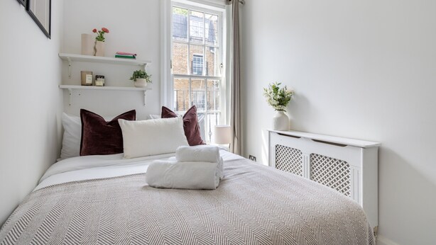 A neatly arranged bedroom with a large window, a radiator, and a shelf with decorative items. - Cosy 2BR in Marylebone Near Baker Street (London)