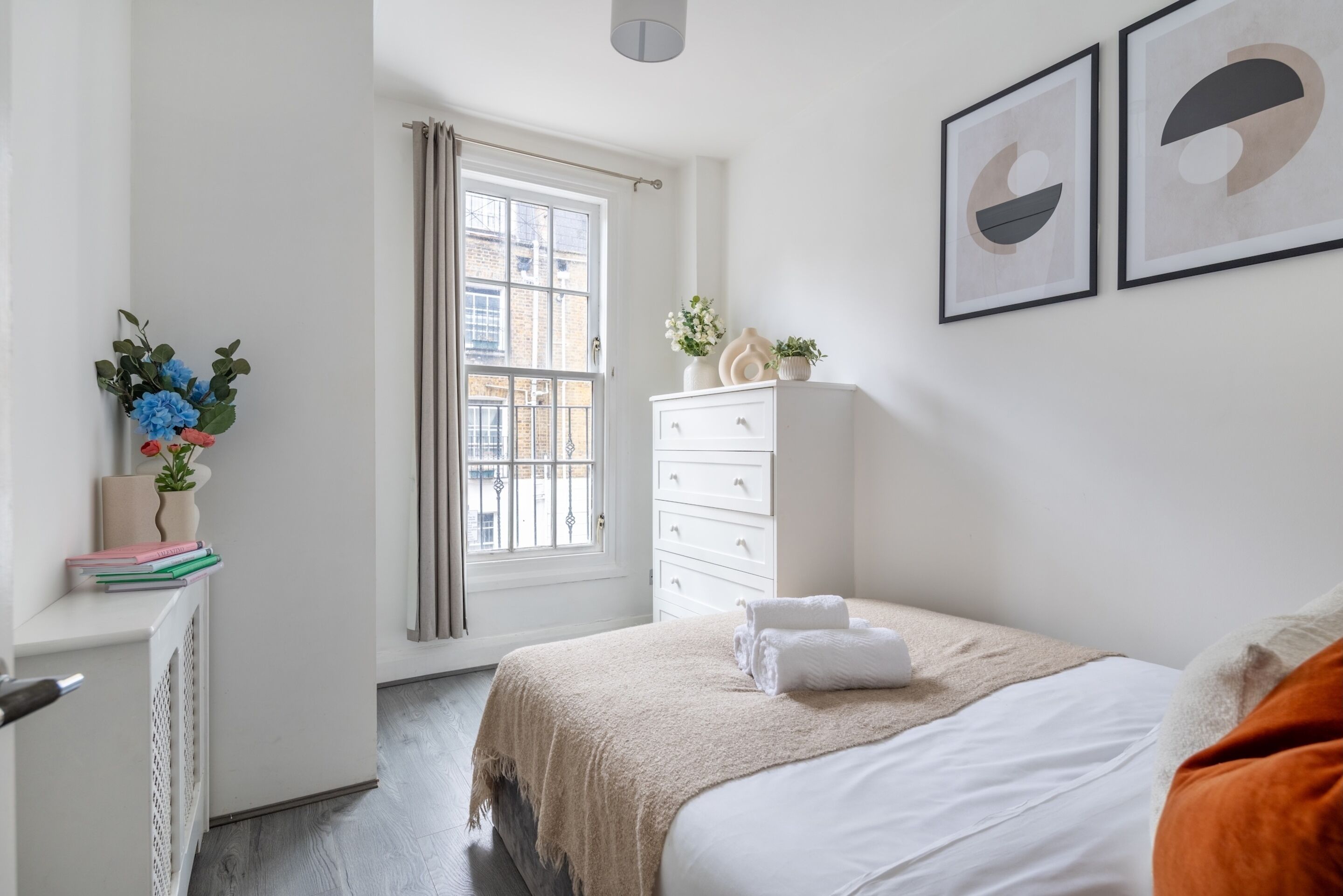 A bedroom with a bed, a dresser, a window with curtains, and two framed artworks on the wall.