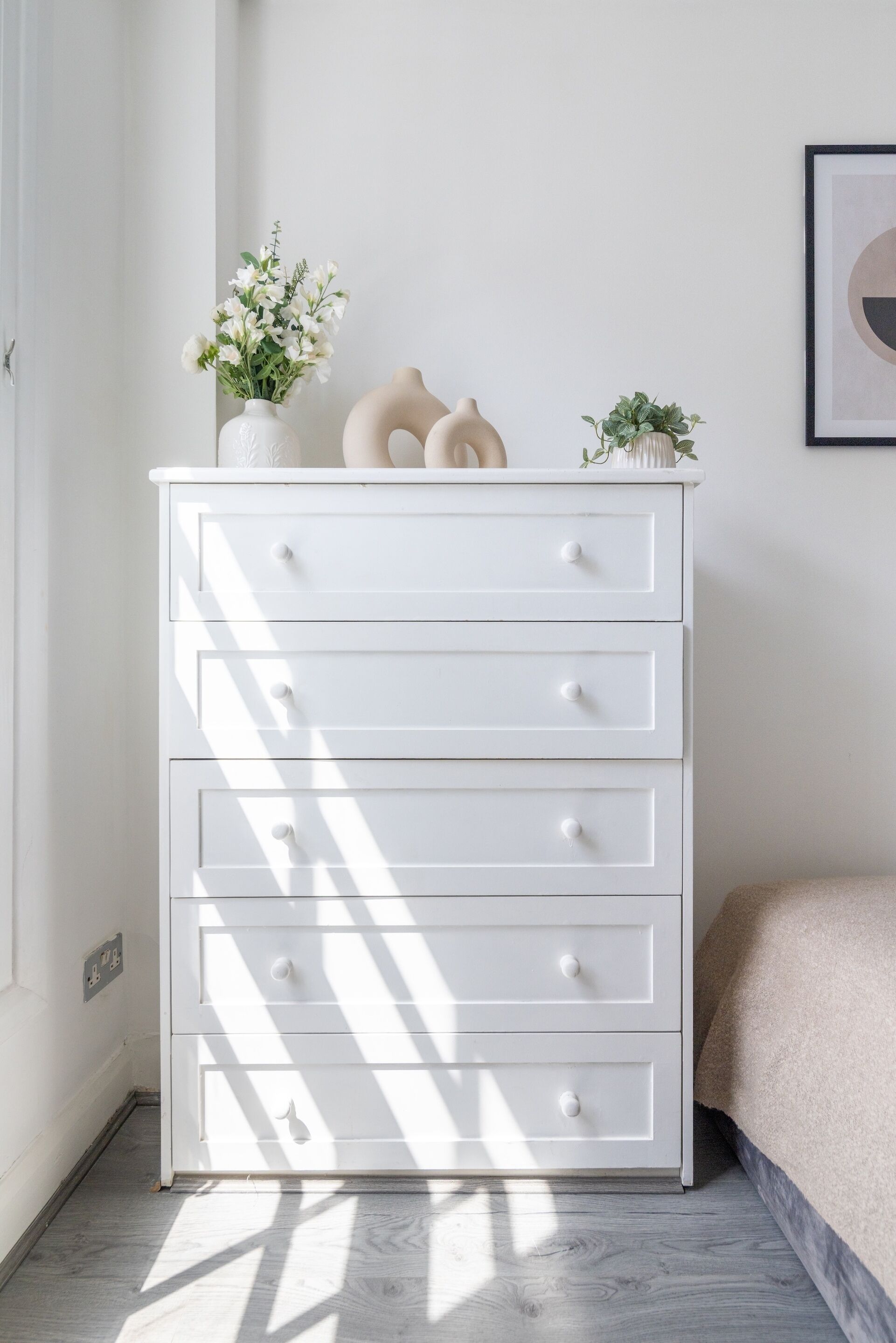 A white dresser with drawers, a vase with flowers, and a framed picture on the wall.