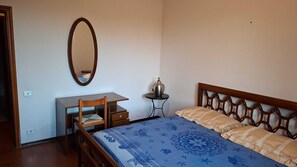 A bedroom with a bed, a desk, a chair, a small table with a lamp, and a mirror on the wall.