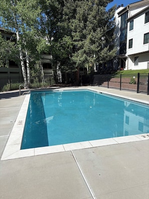 Ein rechteckiger Pool mit glasklar blauem Wasser, umgeben von einer gepflasterten Liegefläche und einem schwarzen Metallzaun.