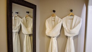 Une salle de bain avec trois peignoirs blancs suspendus à des cintres en bois contre un mur, un grand miroir à cadre foncé et un porte-serviettes avec une serviette.