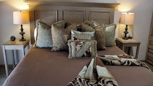A neatly made bed with multiple pillows, two bedside lamps, and a wooden headboard. - The Diamond Suite is our flagship luxury suite. (Fredericksburg)