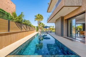 A swimming pool with a wooden deck, a glass wall, and a modern building with a balcony. - Palais Arbaa (Marrakech)
