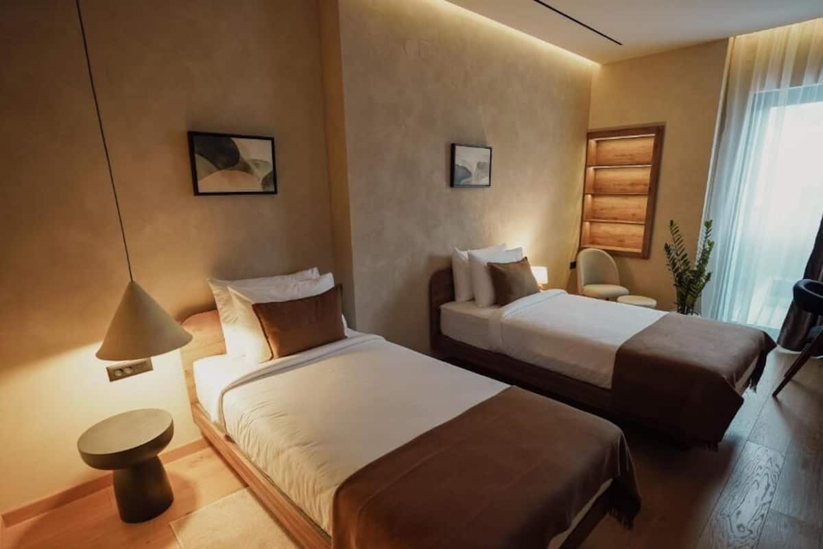 A hotel room with two beds, a bedside table, a lamp, a chair, and a window with curtains.