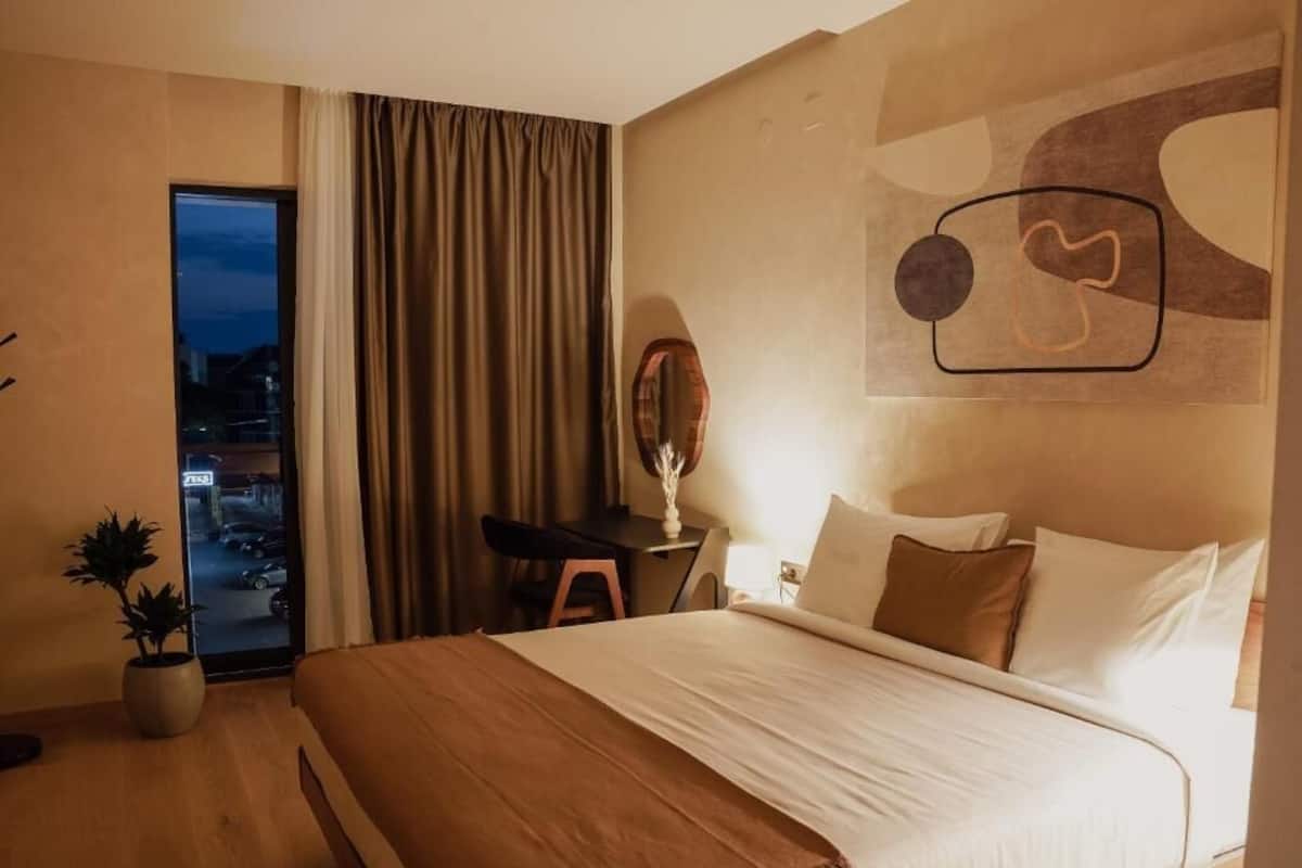 A hotel room with a bed, a desk, a chair, a potted plant, a mirror, and a view through the window.