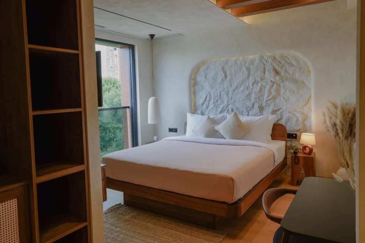 A modern bedroom with a large bed, a wooden headboard, a bedside table with a lamp, and a view of greenery outside.