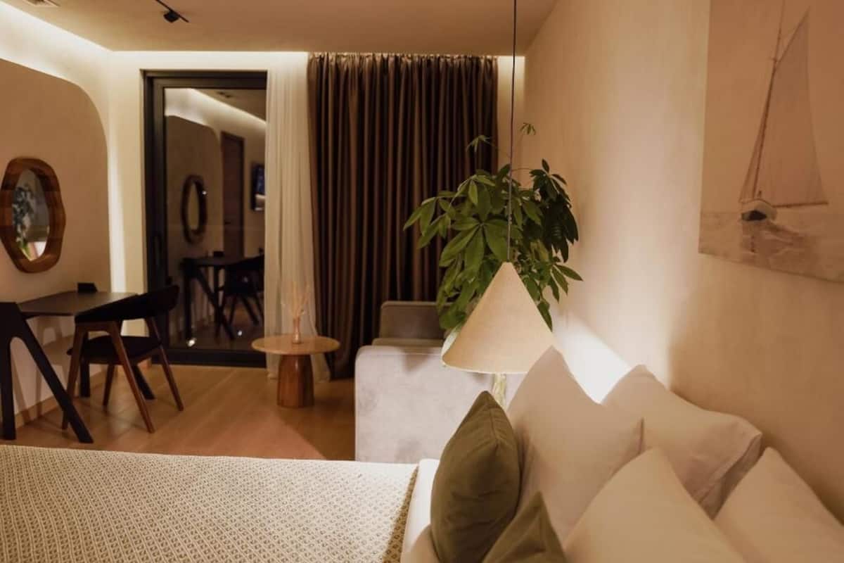 A hotel room with a bed, a desk, a chair, a small table, a lamp, a plant, and a painting on the wall.