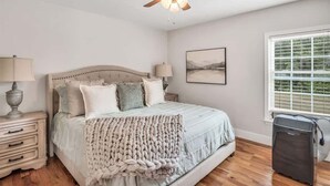 Une chambre à coucher comprenant un lit, des lampes de chevet, un ventilateur de plafond, une fenêtre avec des stores et un tableau accroché au mur.