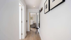 Un pasillo con piso de madera, paredes blancas y una puerta que da a una habitación con un sofá gris y una pequeña mesa negra.