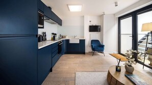Una cocina moderna con armarios azul oscuro, una encimera blanca y suelo de madera. Hay una silla azul, un televisor montado en la pared y una ventana con vistas al exterior.