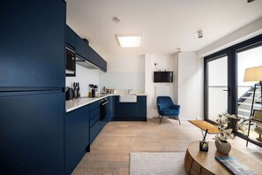 A modern kitchen with dark blue cabinets, a white countertop, and a wooden floor. There is a blue chair, a TV mounted on the wall, and a window with a view of the outside. - 1 Min Walk to Euston Station, Modern Apartment (Greater London)