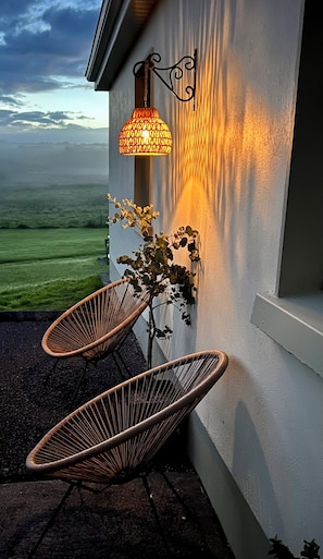 Una silla de mimbre frente a un edificio, con una lámpara de pared y vista al paisaje.