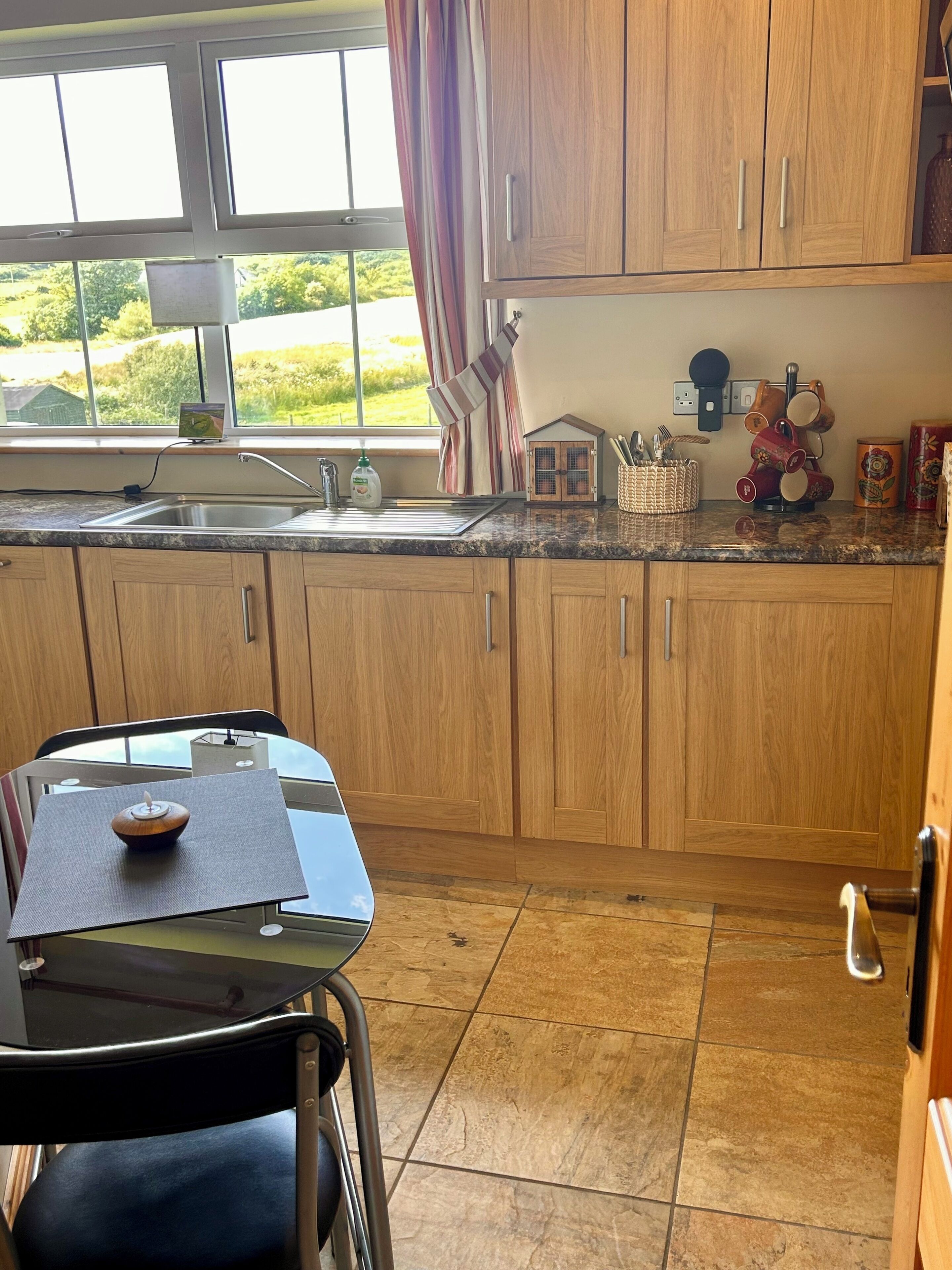Una cocina con armarios de madera, encimera de granito, ventana con cortinas, un pequeño adorno y una vela sobre una mesa.