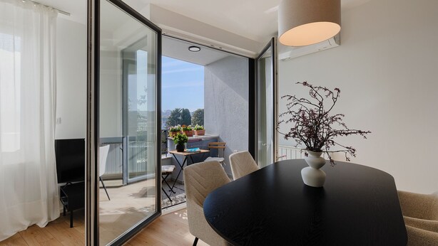 Ein modernes Wohnzimmer mit einem schwarzen Esstisch, einer Vase mit Zweigen und Blick auf einen Balkon mit Blumentöpfen.