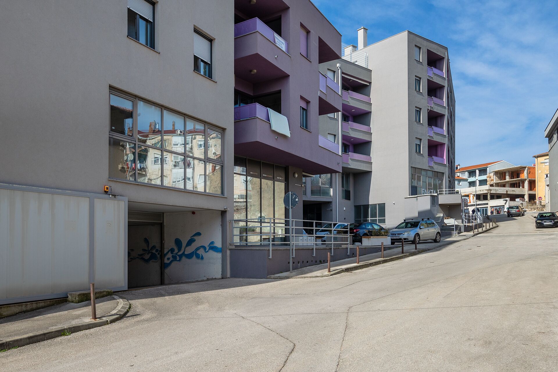A modern multi-story building with balconies, a gated entrance, and parked cars along the street.