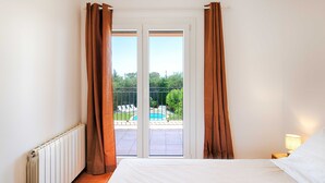 Un dormitorio con cama, mesita de noche, lámpara y vistas a una piscina a través de un ventanal corredera.