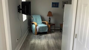 Un couloir étroit avec un plancher en bois, un fauteuil bleu, une petite table avec une lampe et une porte blanche.