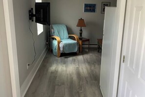 Ein schmaler Korridor mit Holzboden, einem blauen Sessel, einem kleinen Tisch mit einer Lampe und einer weißen Tür.