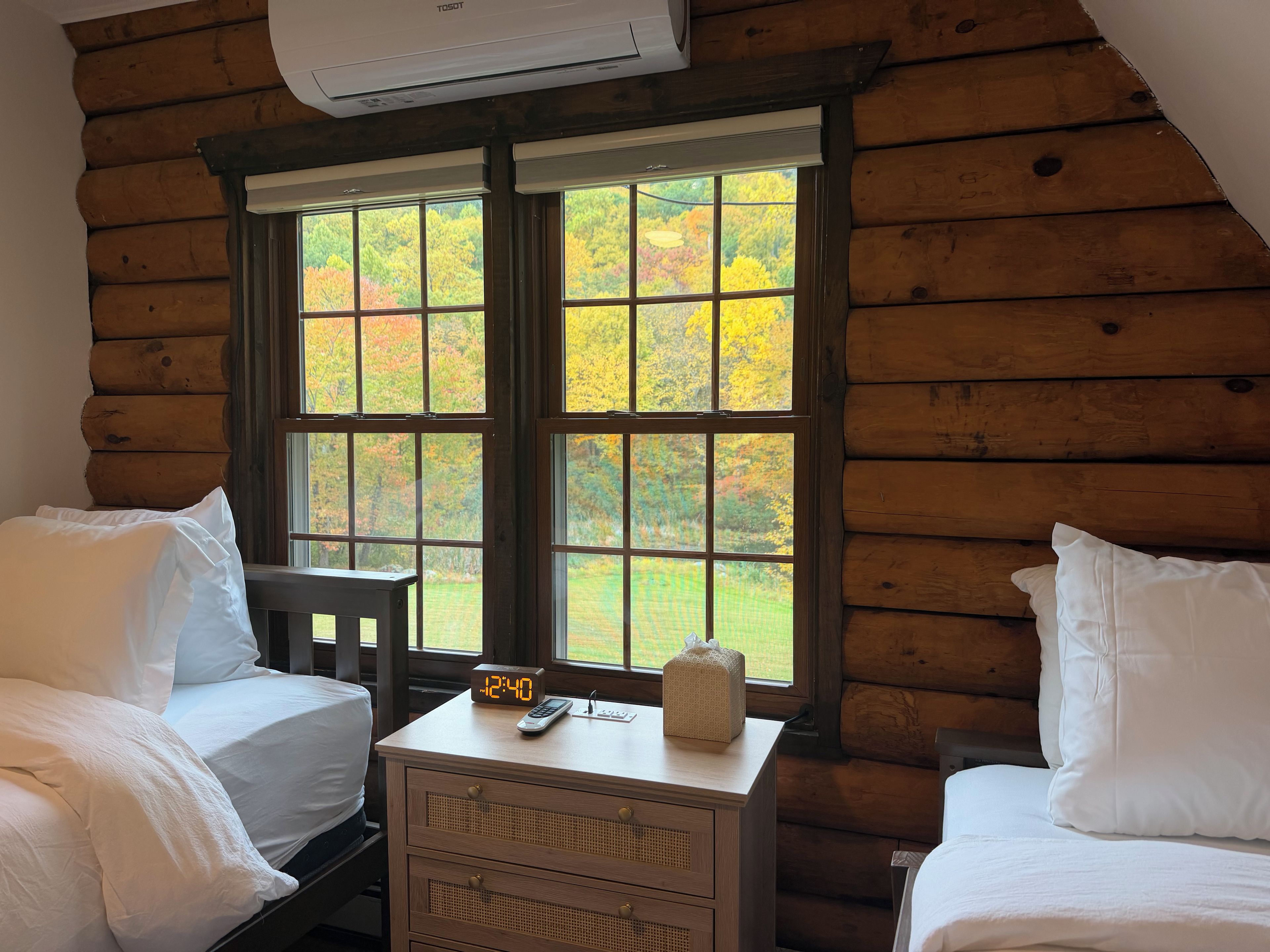 A cabin room with two beds, a nightstand, and a window with a view of autumn foliage.