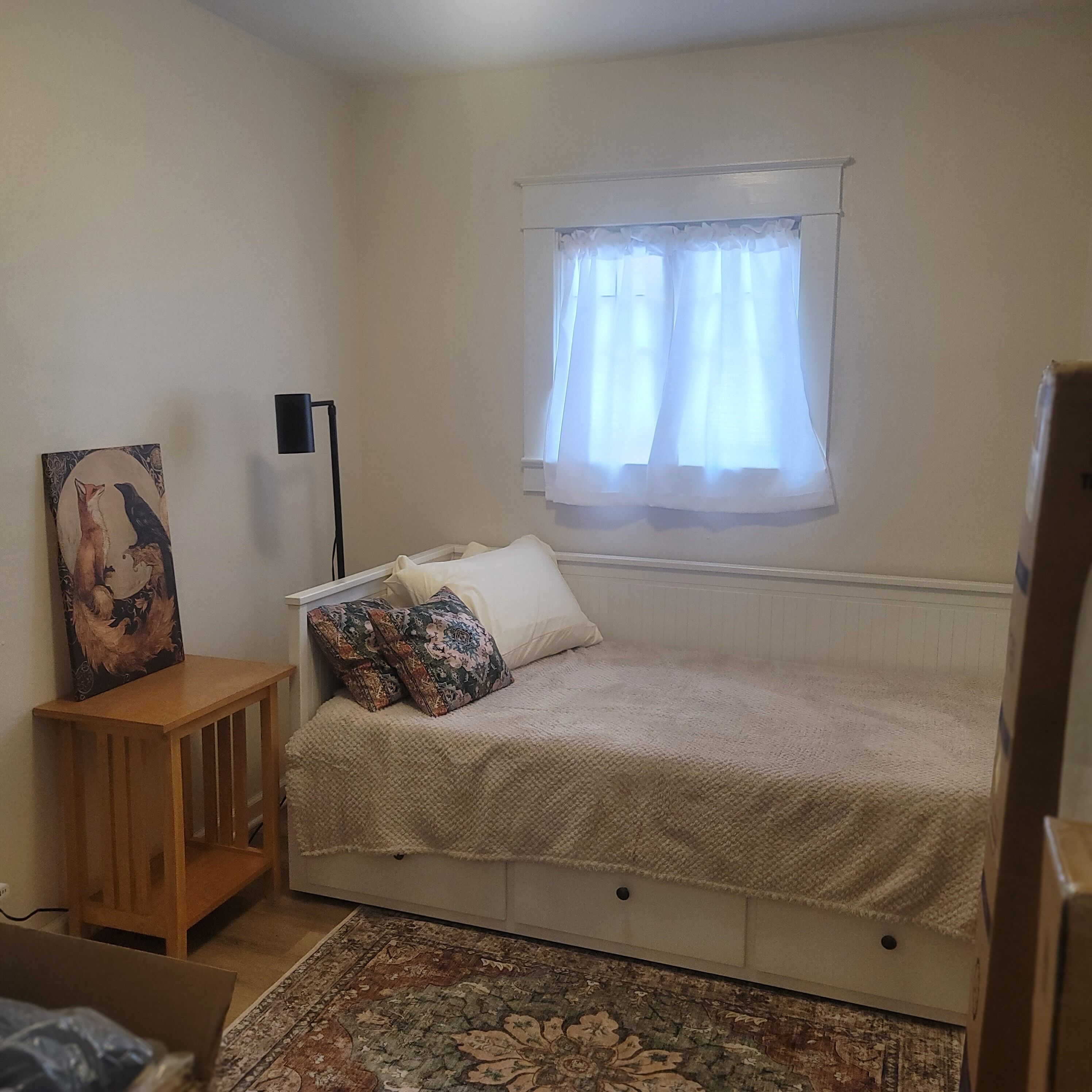 Quarto com cama, mesa de cabeceira, luminária, janela com cortinas e um quadro na parede.