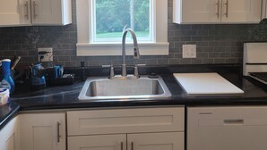 A kitchen with white cabinets, a black countertop, a stainless steel sink, and a window with a view of trees. - Cute, newly remodeled bungalow for 7. (Independence)