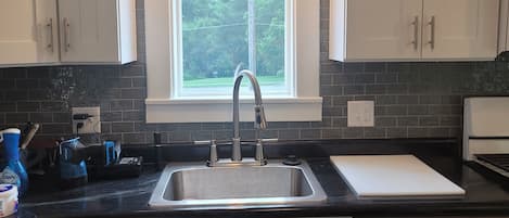 A kitchen with white cabinets, a black countertop, a stainless steel sink, and a window with a view of trees.
