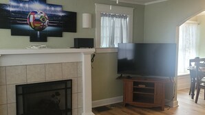 A living room with a fireplace, a flat-screen TV, a ceiling fan, and a patterned rug. - Cute, newly remodeled bungalow for 7. (Independence)