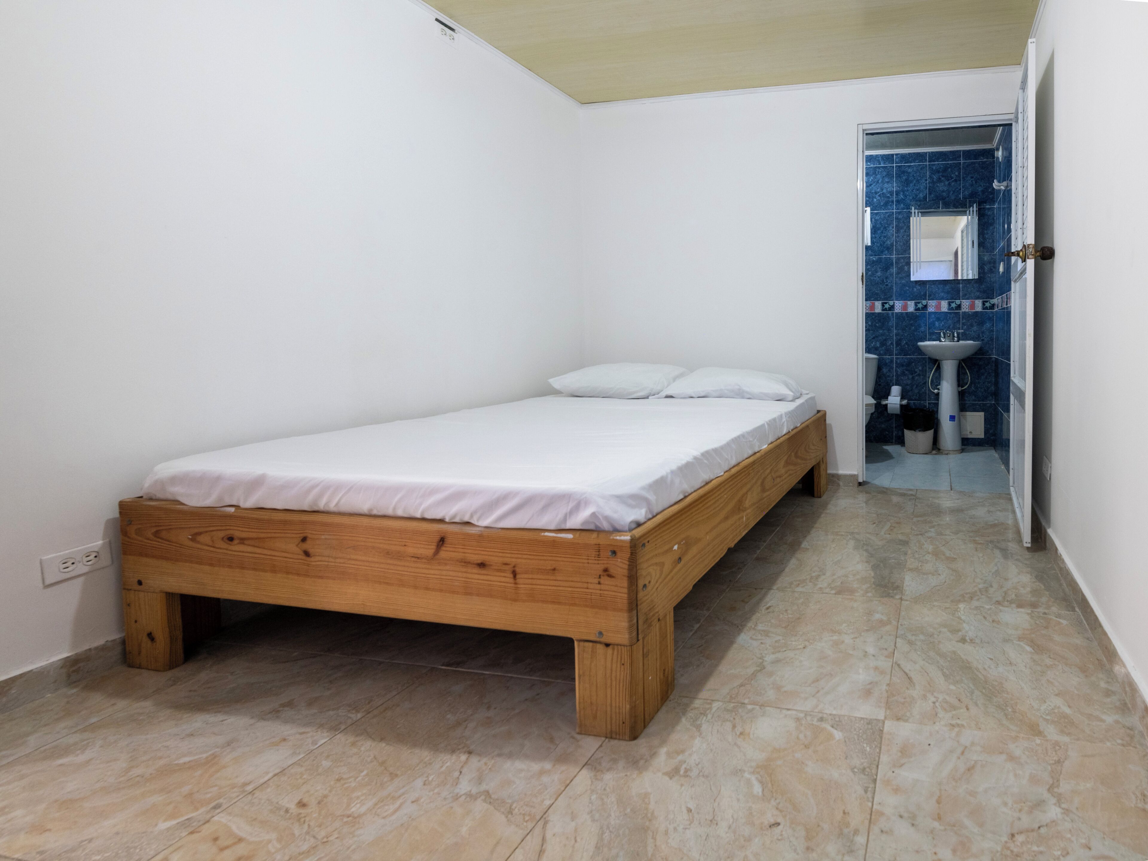 A small, clean room with a single bed, a bathroom with a sink and mirror, and a tiled floor.