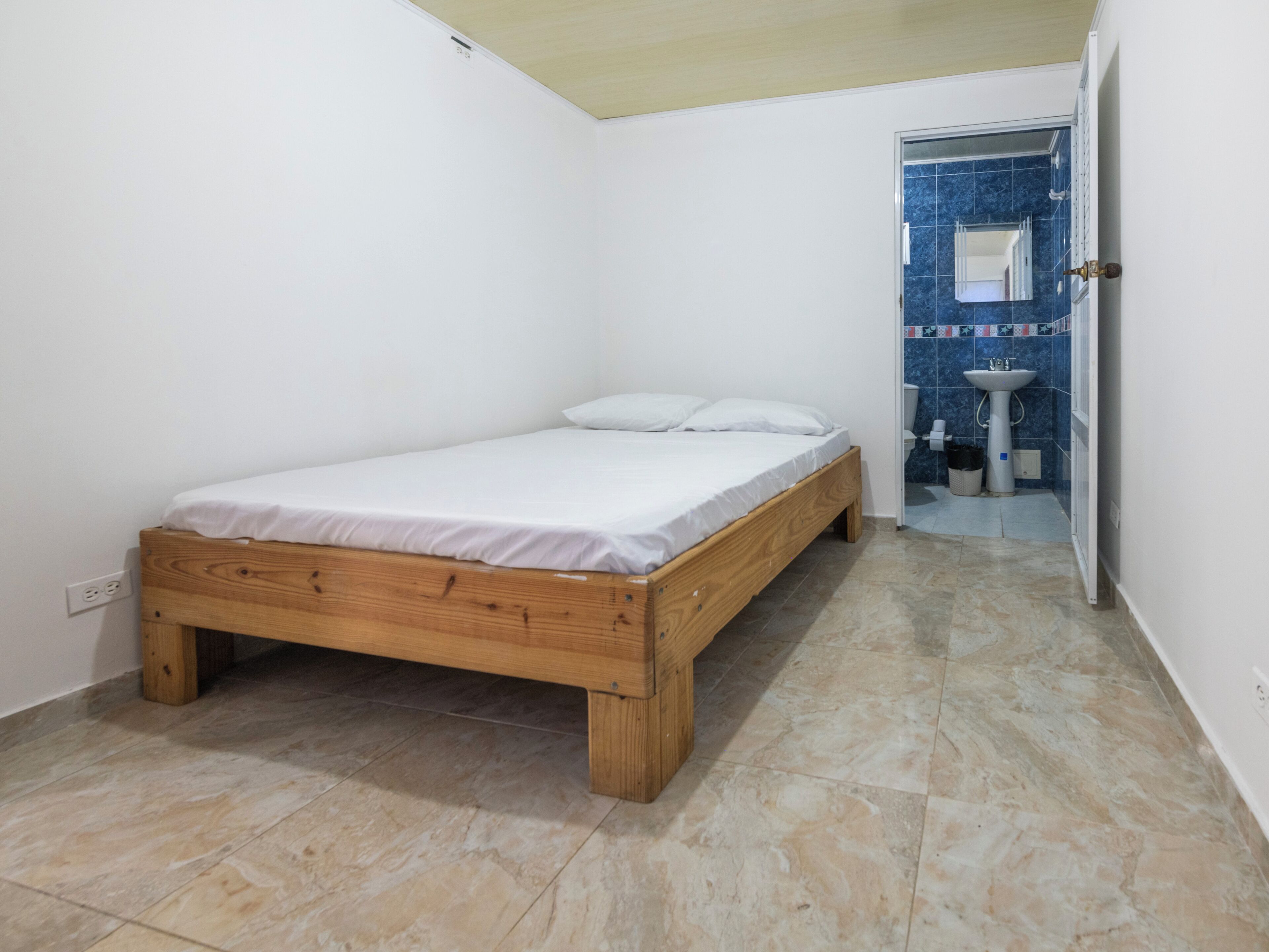 A bedroom with a wooden bed, a white wall, and a bathroom with blue tiles.