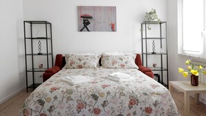 Ein Schlafzimmer mit floralem Bettbezug, zwei schwarzen Metallregalen, einem roten Stuhl, einem kleinen Tisch mit Blumenvase und einem Gemälde an der Wand.