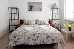 A bedroom with a floral bedspread, two black metal shelves, a red chair, a small table with a vase of flowers, and a painting on the wall. - Very central, bright and multi-accessorised apartment ideal for families. (La Spezia)