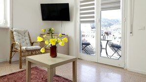 Ein Zimmer mit einem Korbstuhl, einem Tisch mit einer Vase voller gelber Blumen, einem Fernseher an der Wand und Blick durch eine Glastür.