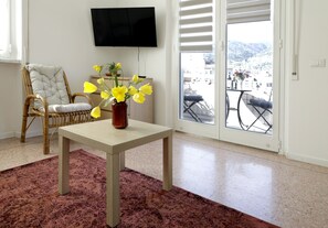 A room with a wicker chair, a table with a vase of yellow flowers, a television on the wall, and a view through a glass door. - Very central, bright and multi-accessorised apartment ideal for families. (La Spezia)