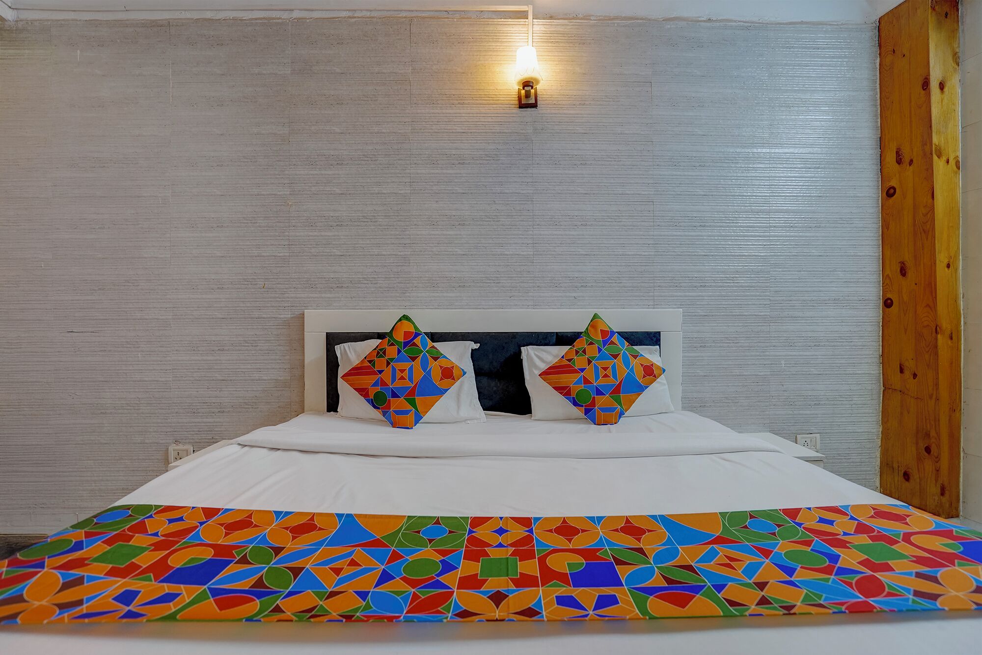 Uma cama com colcha de padrão geométrico, duas almofadas com design semelhante, uma luminária na parede e uma porta de madeira.