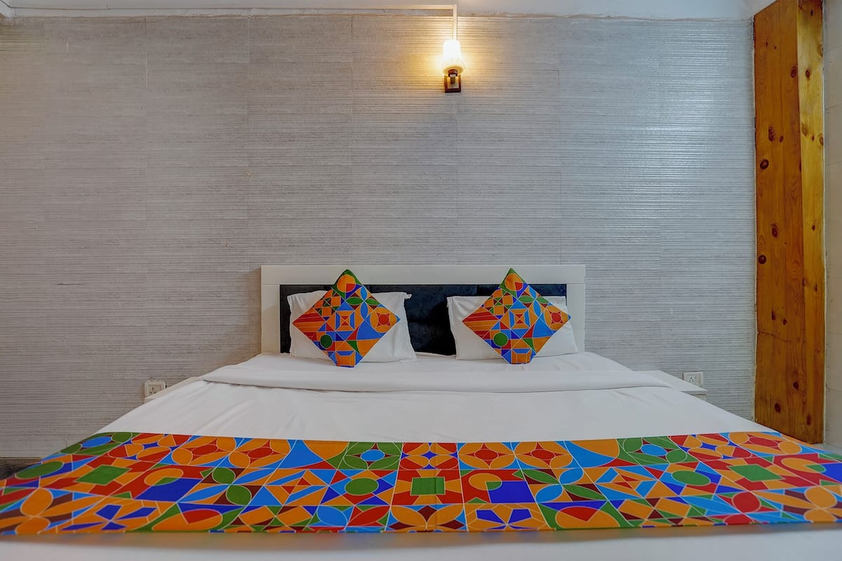 Uma cama com colcha de padrão geométrico, duas almofadas com design semelhante, uma luminária na parede e uma porta de madeira.